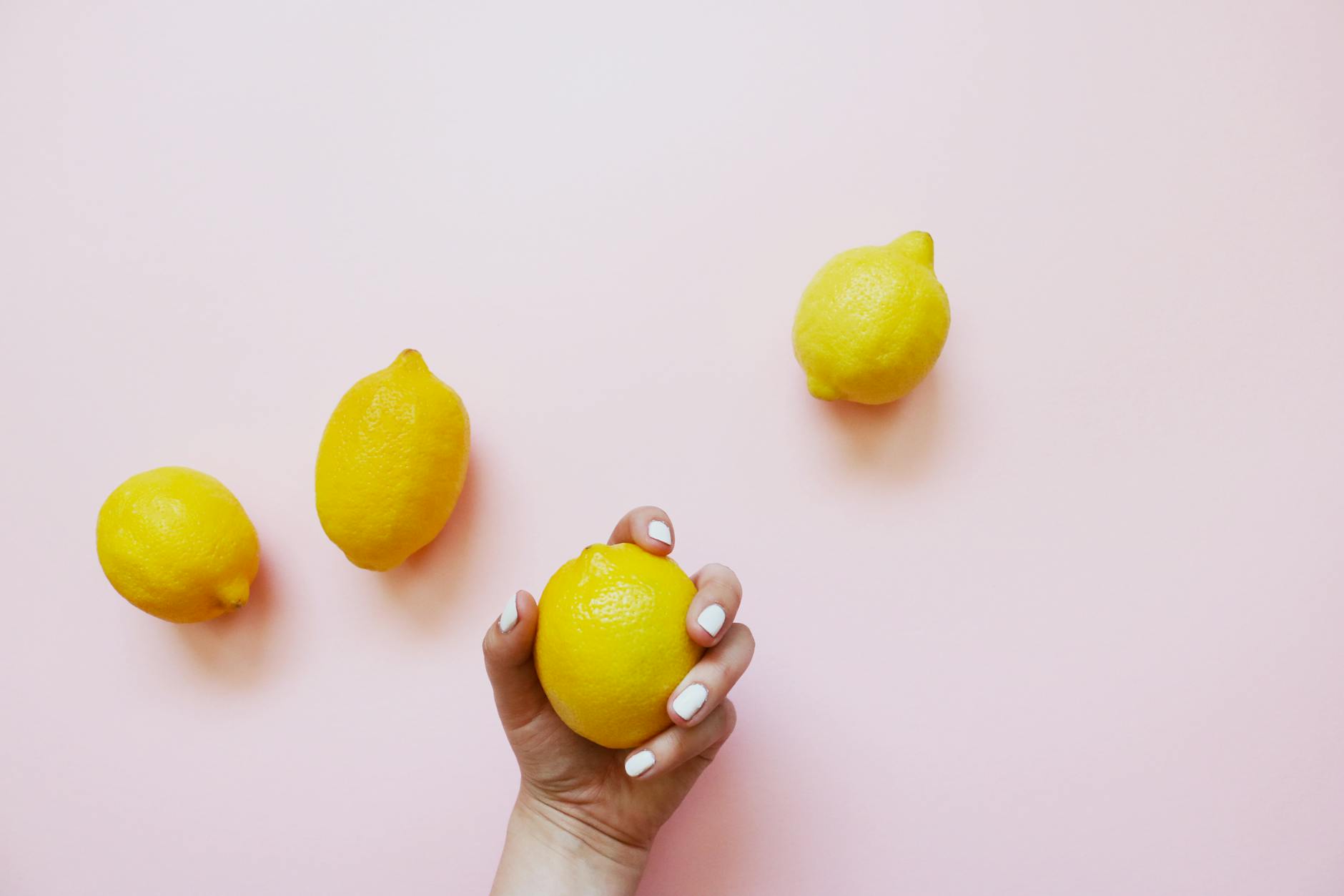 Hand holding a bright lemon on a soft pink background surrounded by fresh lemons